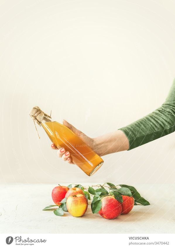 Weibliche Frauen halten eine Flasche mit hausgemachtem Apfelessig in der Hand über einem Tisch mit Äpfeln und grünen Blättern vor weißem Wandhintergrund. Apfelsaison. Konzept für gesundes fermentiertes Essen