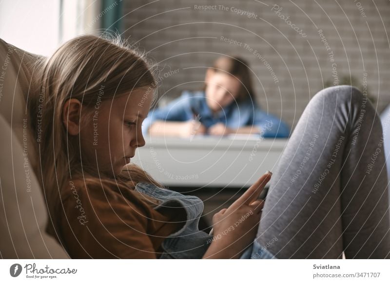 Ein süßes Mädchen spielt am Telefon, während ihre Schwester ihre Hausaufgaben macht. Kommunikation, soziale Distanz während der Quarantäne. Fernunterricht, Online-Bildung