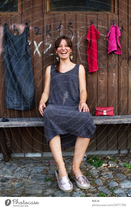 Junge Frau sitzt lachend auf einer Bank, vor einer Garderobe aus Holz, an welcher ein Kleid und Handtücher hängen. Jugendliche Mädchen Mensch Teenager Lifestyle