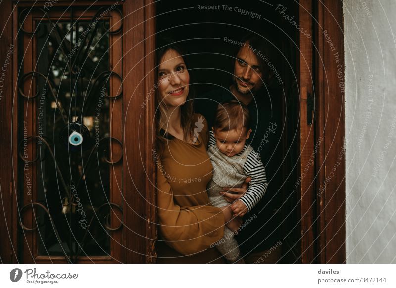 Familienporträt eines jungen Mannes und einer jungen Frau, die ihren kleinen Sohn an der Eingangstür ihres Hauses halten, bei schönem, schwachem Licht. 30s
