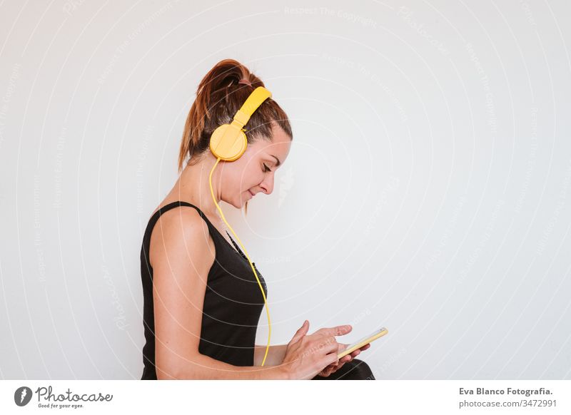 Junge Frau, die mit einem gelben Mobiltelefon und einem Headset Musik hört. Sitzt auf einer Yogamatte und entspannt sich nach dem Sport. Gesunder Lebensstil im Haus