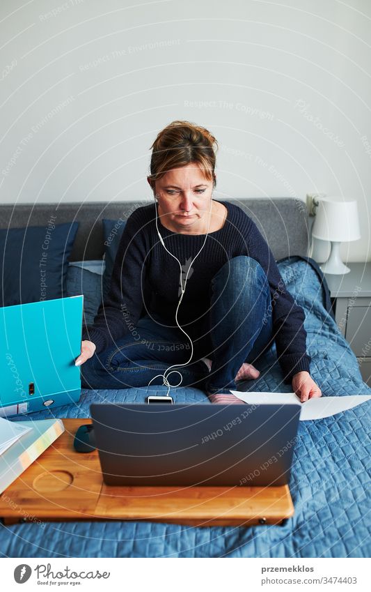 Frau, die während eines Video-Chat-Call-Stream-Online-Kurses per Webinar auf einem Laptop von zu Hause aus ihren Job aus der Ferne erledigt. Frau sitzt im Bett vor dem Computer und schaut mit Kopfhörern und Smartphone auf den Bildschirm