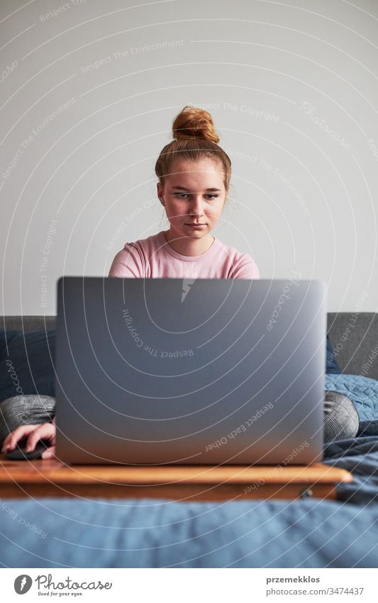 Teenagerin, die während der COVID-19-Quarantäne von zu Hause aus auf ihrem Laptop ferngesteuert Hausaufgaben erledigt. Online-Kursstunden. Lernen aus der Ferne. Mädchen sitzt im Bett vor dem Computer und schaut auf den Bildschirm