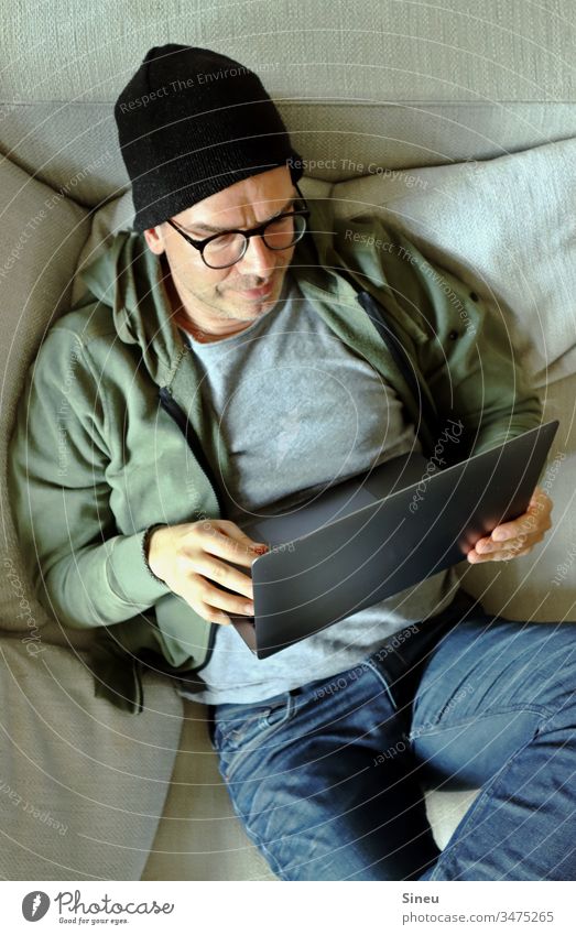 HomeOffice: entspannter Mann liegt auf dem Sofa mit seinem Notebook Homeoffice Arbeitsplatz Mütze Hipster Jeanshose Brille lässig leger selbständig freelancer
