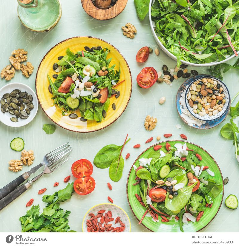 Teller mit grünem Salat auf Küchentischhintergrund mit Gabeln und Zutaten: Nüsse, Samen, junge Blätter, Olivenöldressing. Ansicht von oben. Diät. Sommerliche Küche. Gesunde Hausmannskost