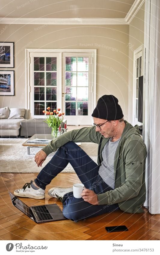 HomeOffice: Entspannter Mann sitzt auf dem Boden vor seinem Notebook und trinkt Kaffee Wohnzimmer Heimarbeitsplatz Ein Mann allein