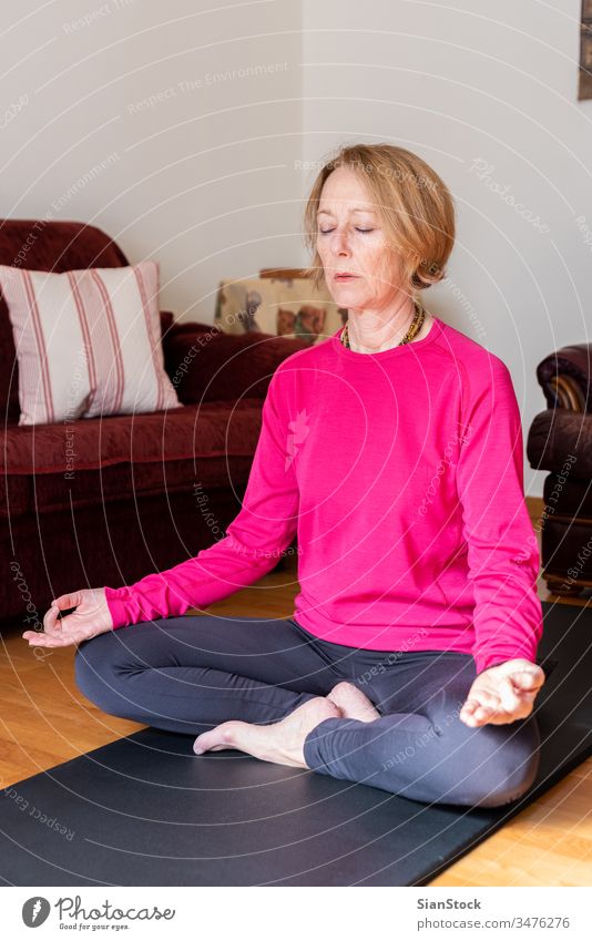 Frau mittleren Alters sitzt im Lotussitz auf einer Yogamatte in ihrem Wohnzimmer. Meditation Lotus-Pose Schönheit Training Europäer trainiert. Flexibilität