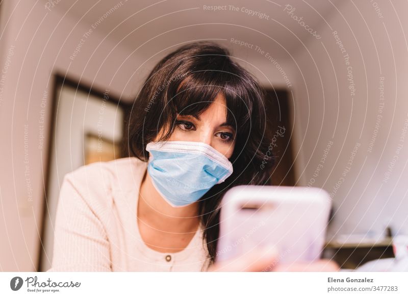 Frau zu Hause mit medizinischer Atemschutzmaske im Gesicht und Mobiltelefon. Chinesisches pandemisches Coronavirus, Virus Covid-19. Quarantäne, Konzept zur Infektionsprävention. Fokus auf ihr Gesicht.