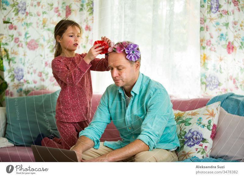 Kind spielt und stört Vater, der fern von zu Hause arbeitet. Kleines Mädchen, das Papas Haare kämmt und eine Frisur macht. Mann sitzt mit Laptop auf der Couch. Familie verbringt Zeit miteinander im Haus.