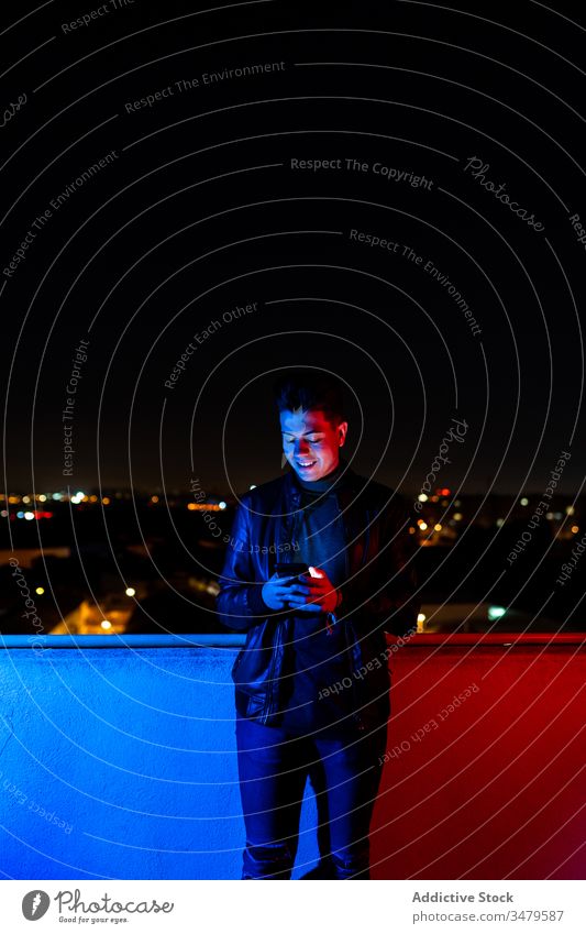 Junger Mann benutzt Smartphone auf beleuchtetem Balkon benutzend Nacht leuchten farbenfroh dunkel modern urban männlich Rotlicht blaues Licht Großstadt Gerät