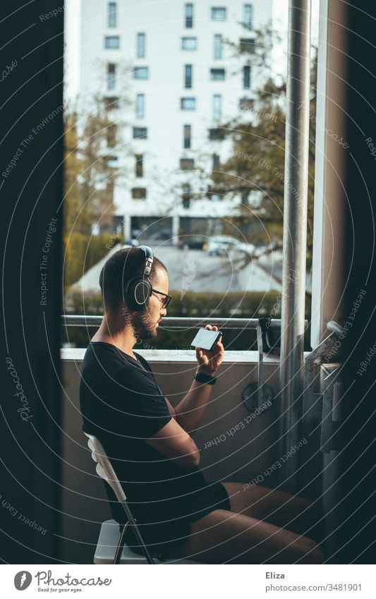Mann mit Kopfhörern sitzt auf dem Balkon in der Abendsonne und sieht etwas auf seinem Smartphone an, Konzept Zuhause bleiben und Social Distancing Medien