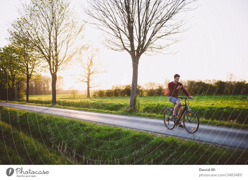 Mann fährt mit dem Handy in der Hand Fahrrad durch die Natur Fahrradfahren Fahrradtour Abgelenkt Sonnenstrahlen Rad Naturerlebnis draußen gutes Wetter Feld