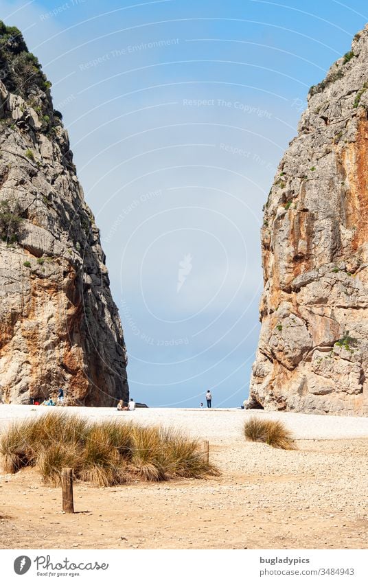 Blick über einen mediterranen Strand zwischen zwei großen Felsen hindurch auf den Himmel. Zwischen den beiden Felswänden sind klein mehrere Personen sitzend und gehend zu erkennen.