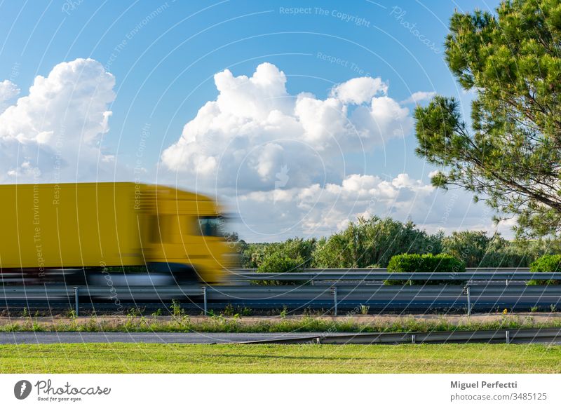 Gelber Lastwagen in Bewegung, der auf der Autobahn neben einem Baum und einem blauen Himmel mit Wolken fährt. Anhänger Ladung Straße Feld Landschaft Verkehr