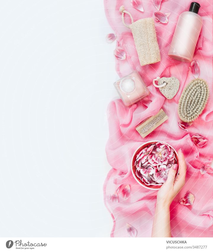Frau hält eine Wasserschüssel mit Blumen auf rosa Handtuch mit verschiedenen umweltfreundlichen Hautpflege- und Schönheitsmitteln: Pinsel, Schwamm, Bimsstein und Kosmetikprodukte auf weißem Hintergrund. Ansicht von oben. Flach liegend