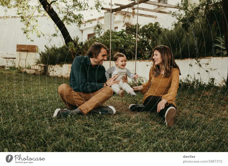 Vater und Mutter schaukeln ihr Baby, das auf dem grünen Gras im Park sitzt. Das Kind lacht glücklich. Aufregung Genuss Lächeln Sitzen Pflege unschuldig Liebe