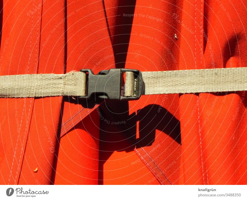 brauner Spanngurt mit schwarzer Schließe hält einen roten Sonnenschirm zusammen Gurt Schutz zusammenhalten Schatten Wetterschutz Baumwollstoff Stoff knallrot