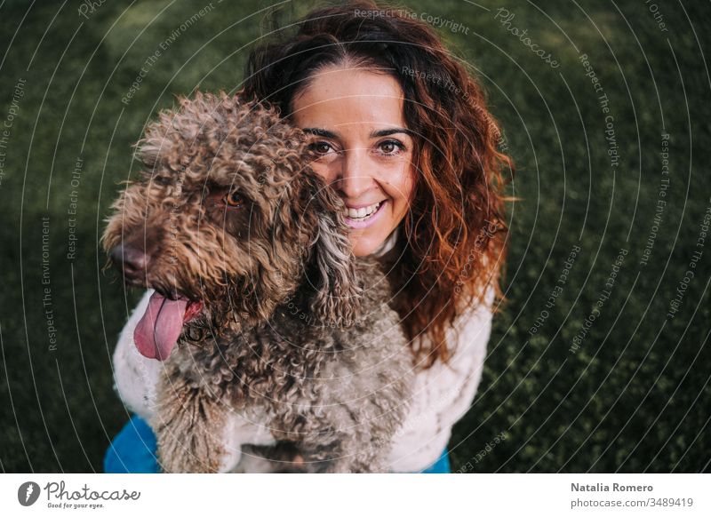 Eine schöne Frau ist mit ihrem Hund auf der Wiese. Die Besitzerin umarmt ihr Haustier, während sie in die Kamera schaut. Sie genießen einen Tag im Park. Das Haustier ist ein spanischer Wasserhund mit braunem Fell.