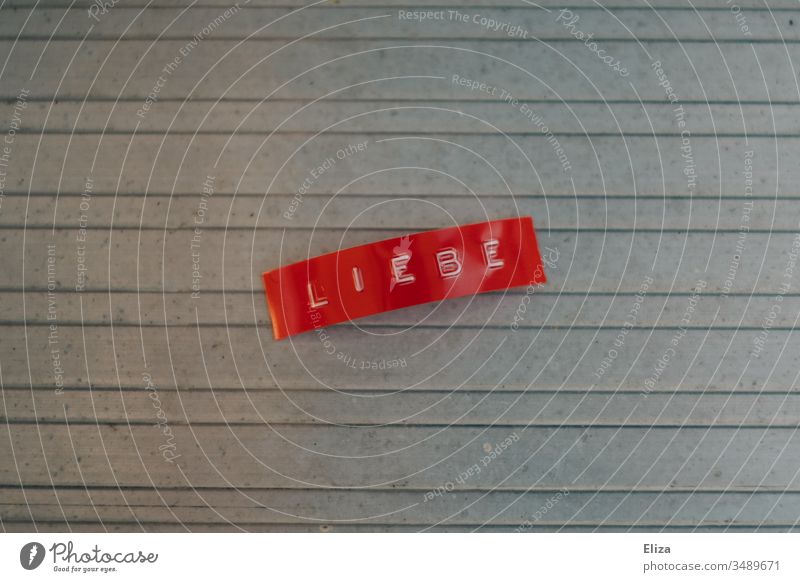Ein rotes Etikett auf dem das Wort Liebe geschrieben steht Buchstaben Schriftzeichen Menschenleer grau Text Gefühle Hintergrund neutral Metall linien