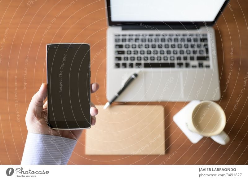 Junger Geschäftsmann hält in der Hand ein Smartphone und in einem unkonzentrierten Hintergrund einen Holzschreibtisch mit einem Laptop, einem Notizbuch und einer Tasse Kaffee.