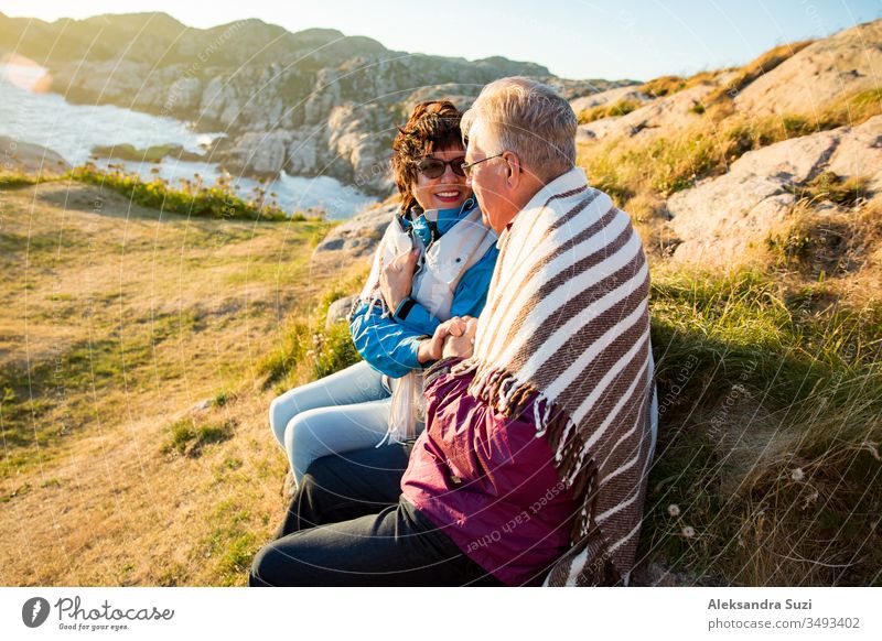 Liebevolles, reifes Paar beim Wandern, auf einer windigen Felsspitze sitzend, auf Erkundungstour. Aktiver reifer Mann und Frau, in eine Decke gewickelt, sich umarmend und glücklich lächelnd. Panoramablick auf Meer und Berge. Norwegen, Lindesnes.