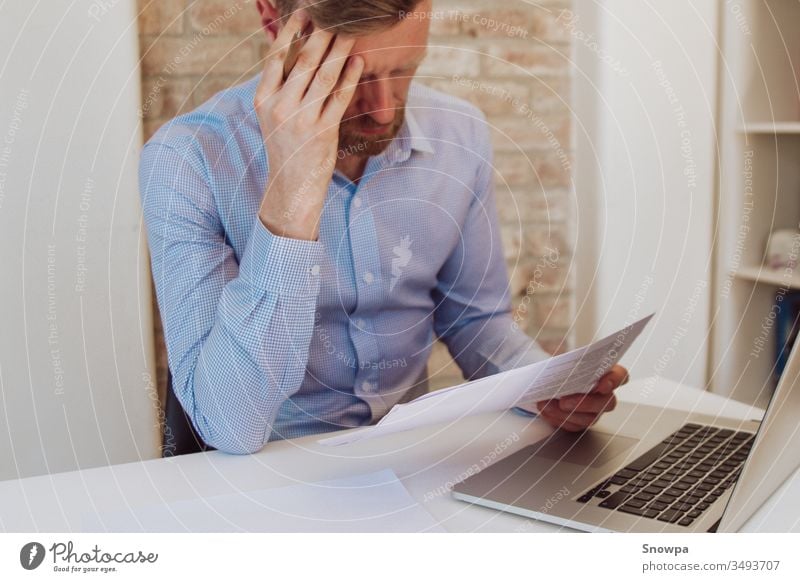 Mann mit Laptop am Schreibtisch sitzend und in hellblauem Hemd Papiere lesend. Heller Arbeitsbereich, Geschäftsraum. Arbeitsumgebung. Backsteinmauer-Hintergrund.