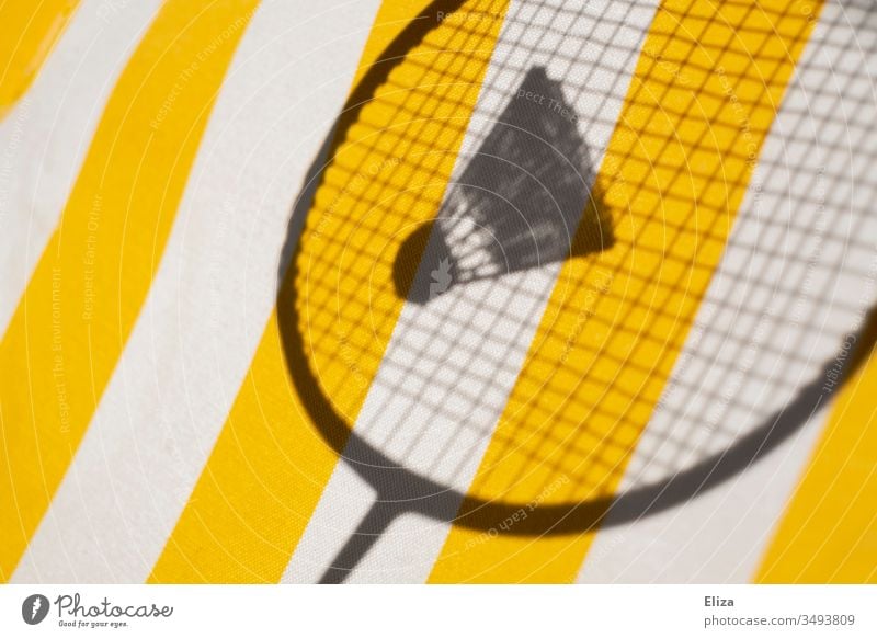 Der Schatten eines Federballschlägers mit einem Federball vor gestreiftem gelben Hintergrund; Badminton Badmmintonschläger Sport Ballsport Freizeit & Hobby