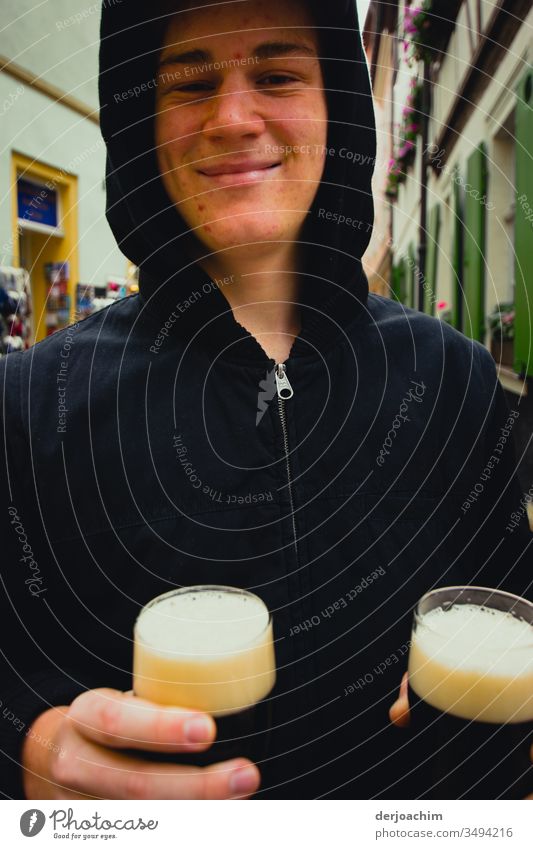Junger Mann hält zwei Gläser mit dunklem  Bier auf der Straße  ( Rauchbier ) in der Hand und lächelt. Der Weiße Schaum  in den Gläsern ist gut an seiner dunklen Jacke zu erkennen.