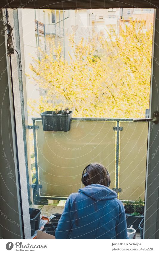 junge Frau sitzt mit Kopfhörern auf dem Balkon zum Hinterhof und pflanzt Blumen und Kräuter Ausblick Balconing Balkonbepflanzung Hobby Hoffnung Musik hören Saat