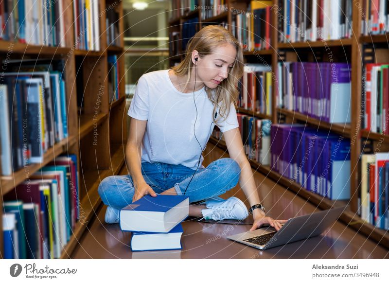 Junges Mädchen, das in der traditionellen Bibliothek am Boden in Bücherregalen sitzt und Bücher liest. Lächelnde und lachende Studentin, die mit dem Laptop arbeitet, lernt. Hochschulbildung.  Studentisches Leben