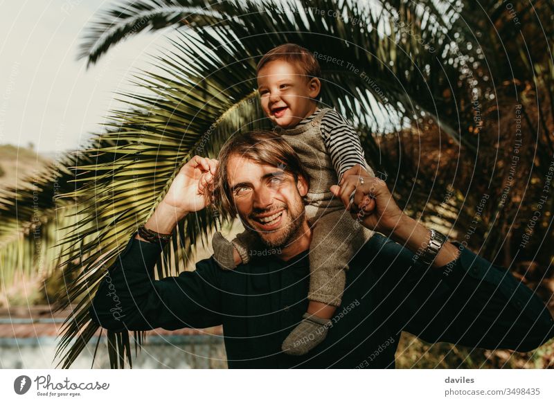 Lachender Vater, der seinen Sohn huckepack im Freien mit Sonnenlicht im Gesicht reitet. lieblich Lächeln weiß Spielen tragen Schultern gutaussehend freudig