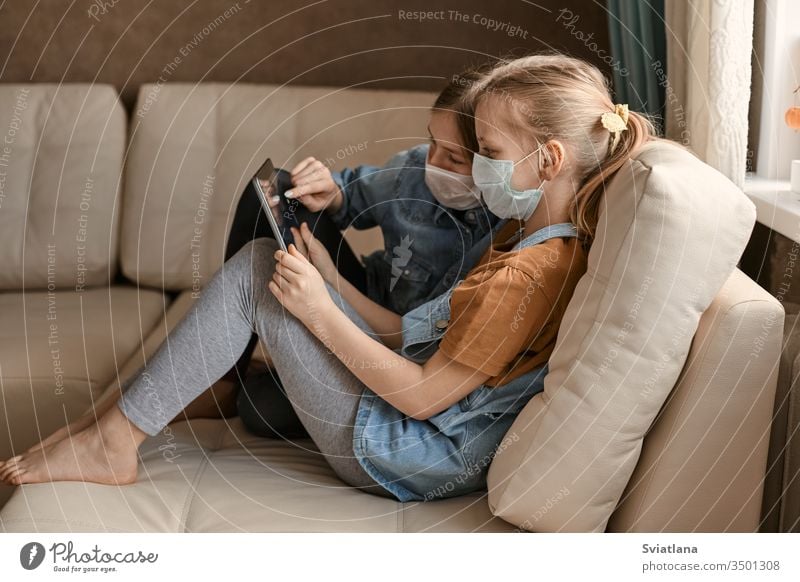 Zwei süße Mädchen mit medizinischen Masken sitzen mit einer Tablette auf dem Sofa. Schulmädchen machen ihre Hausaufgaben. Fernunterricht während der COVID-19-Quarantäne.