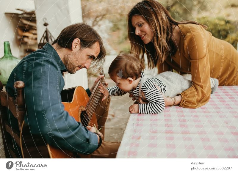 Ein einjähriges Baby, das die Gitarre seines Vaters berührt, während es auf dem heimischen Hof spielt. Die Mutter hält das Baby. klein Windstille Garten lässig