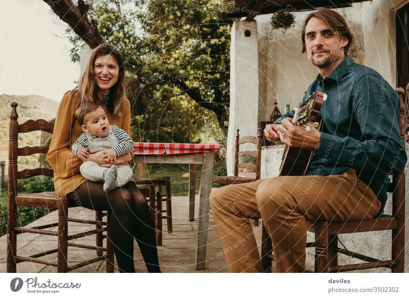Schönes Paar mit ihrem Kleinkind, das gerne auf dem Hof auf dem Land sitzt. Mann spielt Gitarre. Frau hält das Baby. Sie schauen in die Kamera. klein Windstille