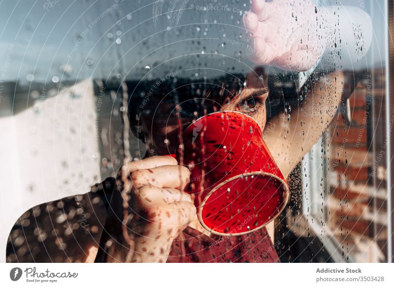 Nachdenkliche Frau trinkt Kaffee am Fenster traurig Depression trinken Selbstisolierung heimwärts unglücklich einsam COVID19 Melancholie jung nass Regen