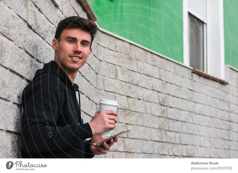 Fröhlicher Mann mit Smartphone und Getränk zum Ausruhen auf der Straße benutzend Großstadt fettarm Wand trinken zum Mitnehmen positiv männlich Gebäude jung