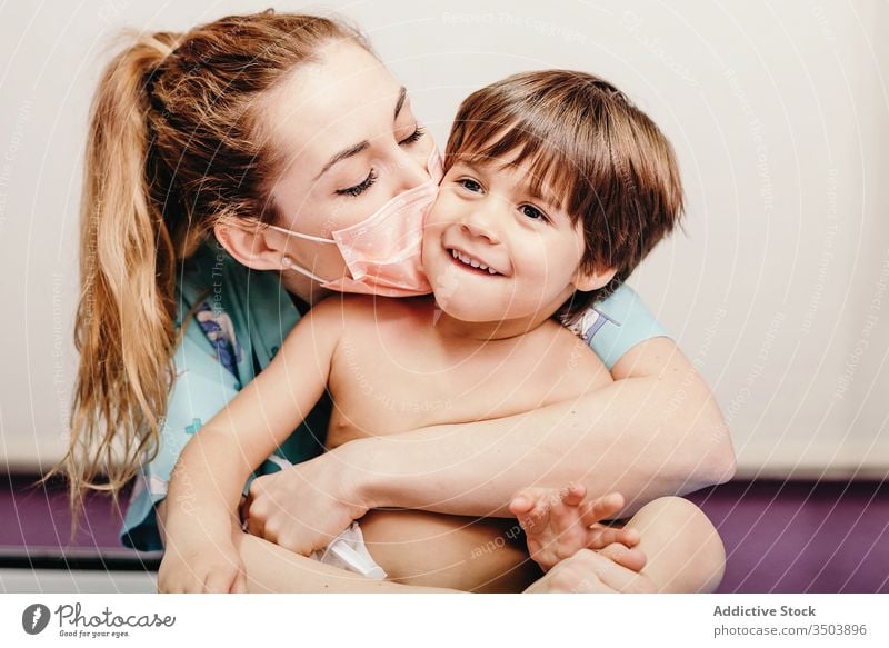 Glückliche Frau mit medizinischer Maske küsst Kind Mundschutz Kuss geduldig Klinik Art Liebe Umarmung verhindern behüten Virus Zusammensein heiter jung niedlich