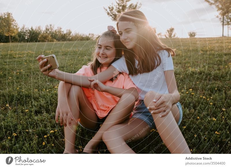 Bezaubernde Schwestern, die in einem Park sitzen und sich ein Selfie Kind Natur Zusammensein Glück Umarmung Gras Sommer Mädchen Landschaft heiter Teenager grün