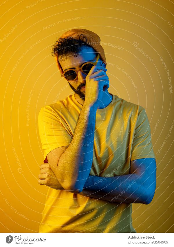 Selbstbewusster bärtiger Mann unter gelber Beleuchtung modern lässig Stil Gesicht berühren ernst Vollbart gelbes Licht hell Outfit männlich trendy Hut T-Shirt