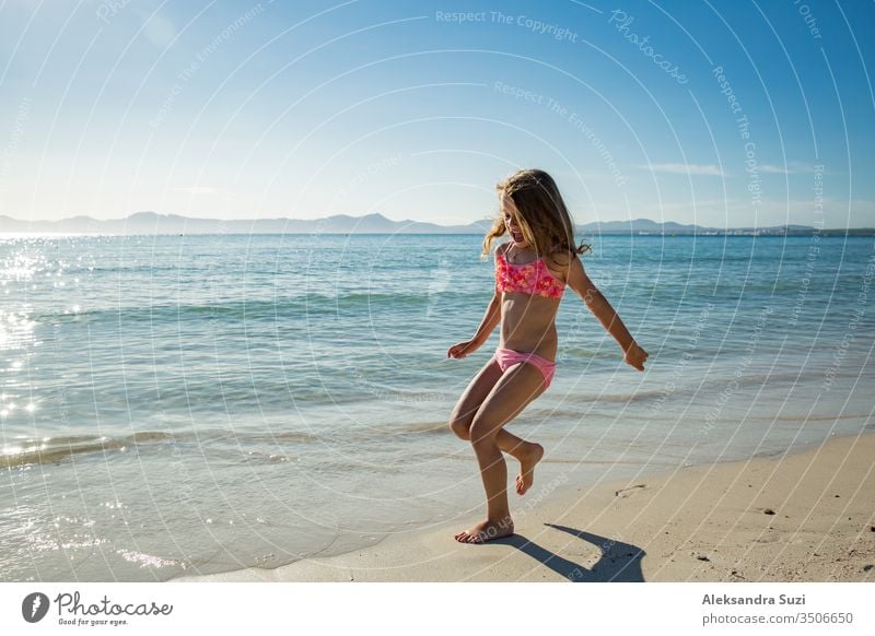 Süßes glückliches kleines Mädchen, das im Badeanzug am Strand entlang läuft und über Wellen springt. Wunderschöner sonniger Sommertag, blaues Meer, malerische Landschaft. Mallorca, Spanien