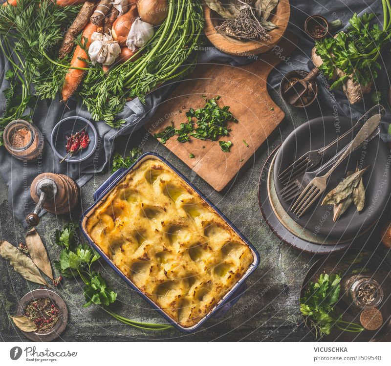 Gebackenes Kartoffelpüree in Kasserolle mit Löffel auf dunklem rustikalem Küchentischhintergrund mit Zutaten und alten Utensilien. Ansicht von oben. Schmackhafte Hausmannskost