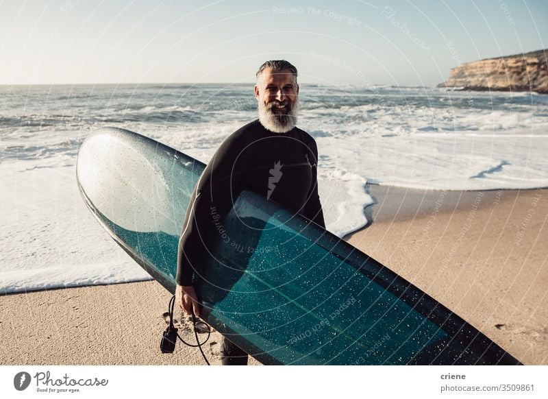 Lächelnder älterer Mann mit Surfbrett am Strand, der in die Kamera schaut Glück Senior Männer Urlaub Surfen Erwachsener graue Haare Lifestyle Freude Sport Hobby