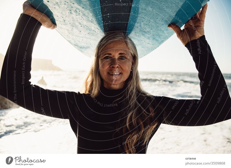 Lächelnde ältere Frau trägt Surfbrett am Strand und schaut in die Kamera Senior Frauen Urlaub Surfen Erwachsener graue Haare Lifestyle Freude Kaukasier Sport