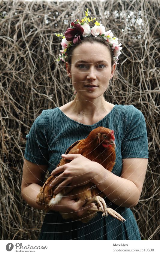 Junge Frau mit Blumenkranz im Haar steht im Hühnerauslauf hält braunes Huhn im Arm Zentralperspektive Schwache Tiefenschärfe Tag Außenaufnahme Farbfoto Idylle