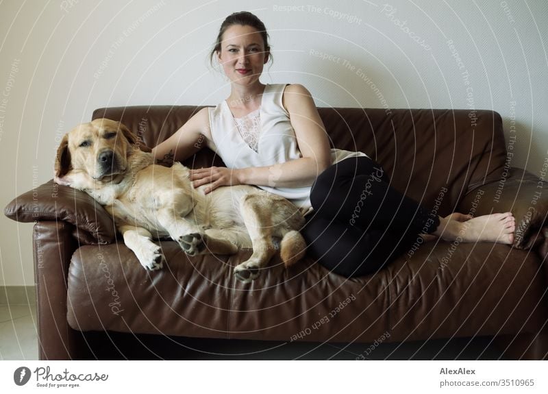 Blonder Labrador und junge Frau sitzen auf einer braunen Ledercouch Haustier Hund blond niedlich liegt sitzt Jugendliche 18-30 Jahre Couch innenaufnahme schlank