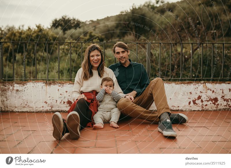 Fröhliches Familienporträt mit Mann und Frau, die mit ihrem Baby auf dem Boden auf der heimischen Terrasse sitzen. wirklich authentisch alternativ patio heiter