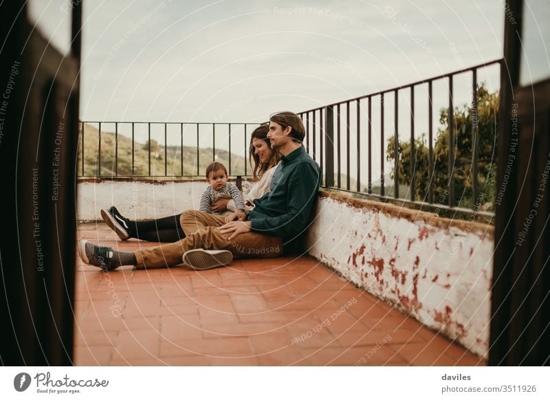 Glückliche Familie mit Mann und Frau, die mit ihrem Baby auf der heimischen Terrasse auf dem Boden sitzen, mit entspanntem Ausdruck. wirklich authentisch