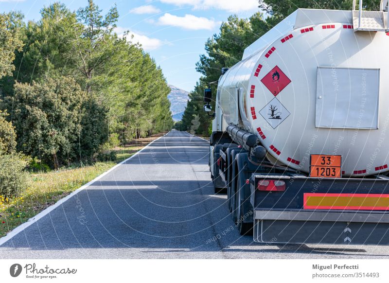 Gefahrzettel für brennbare und umweltschädliche Flüssigkeiten auf einem Tankwagen, der auf einer schmalen, geraden, von Bäumen gesäumten Straße fährt.