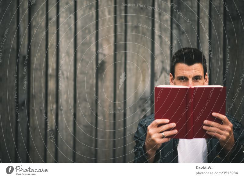 Mann hält vor einer Holzwand ein Buch in den Händen und schaut in die Kamera lesen Bildung gebeildet lernen Lesestoff Buchseite Leser freude Spaß Literatur