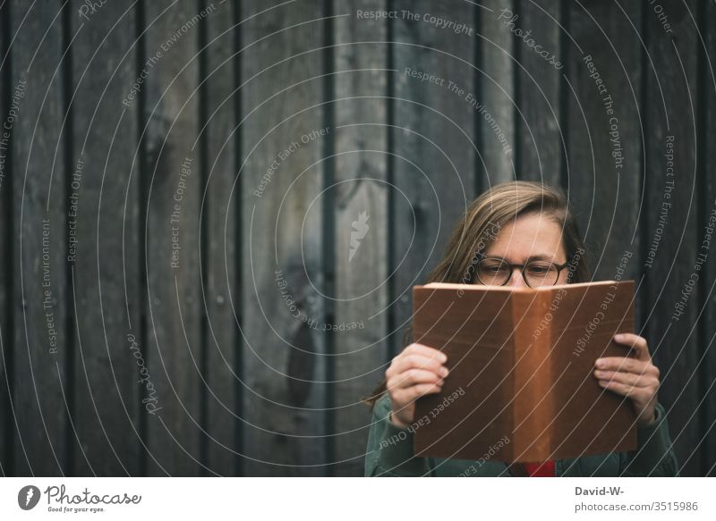 junge Frau mit Brille liest draußen vor einer rustikalen Holzwand ein Buch lesen Bücher dunkel lesend gebildet Bildung interessiert schlau wissensdurstig